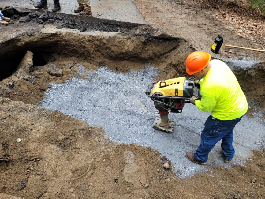 Highway department employee tamping crushed stone backfill at culvert project.